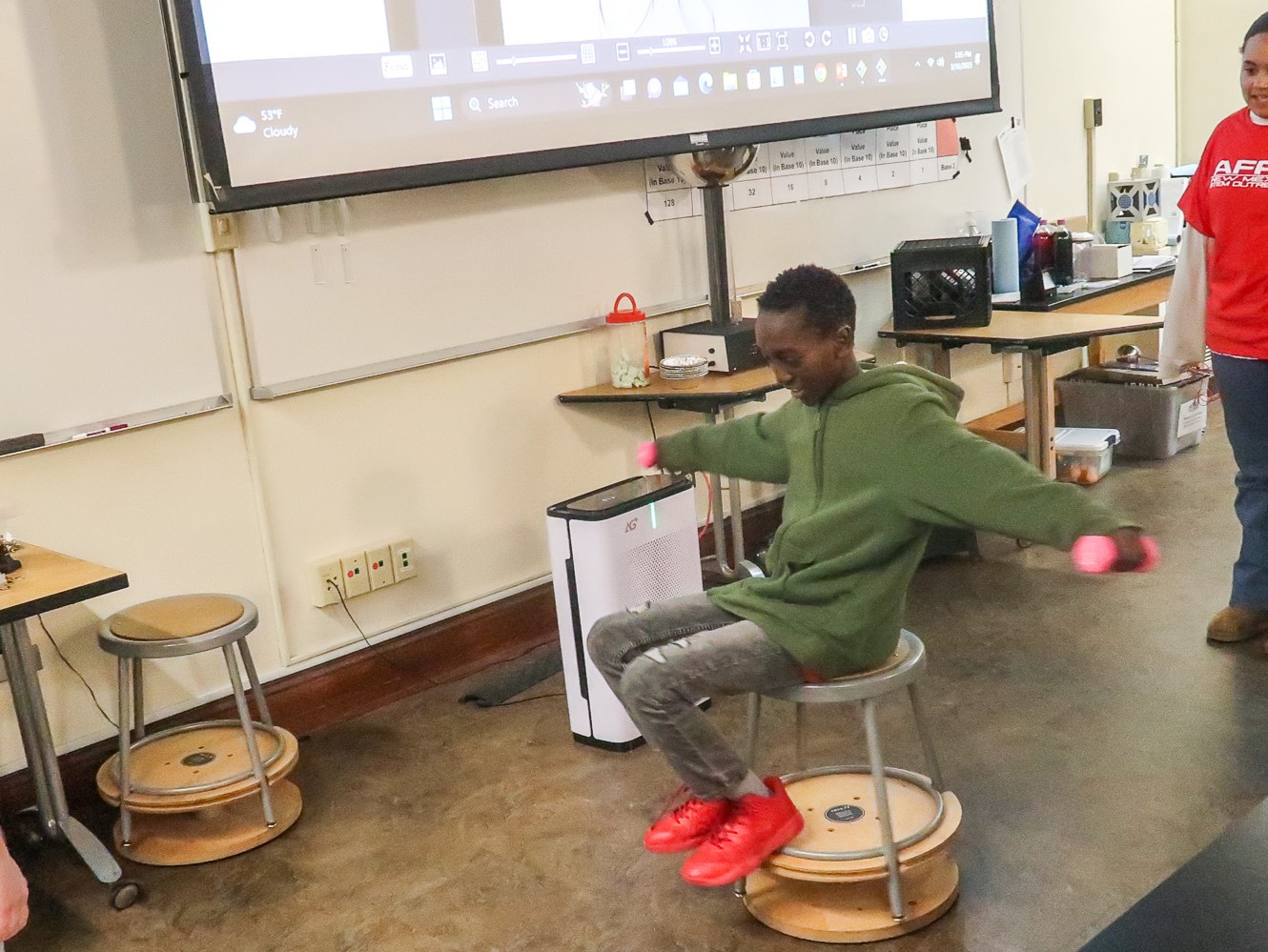 A student sits on a stool with arms outstretched holding small weights, while he spins